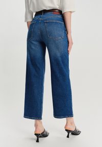 Persona che indossa jeans cropped a gamba larga blu scuro, una maglietta bianca, e mules con tacco neri, mostrata da dietro su uno sfondo semplice.