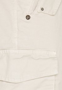 Krémová bavlněná tkanina s jemnou žebrovanou strukturou, obsahující kovový patent a náprsní kapsu se klopami. Čisté linie a detaily šití.