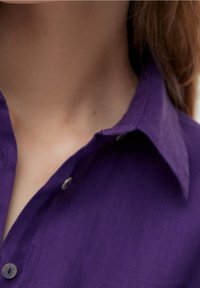 Gros plan sur le cou et le haut de la poitrine d'une personne portant une chemise violette à col avec deux boutons visibles.