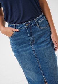 Denim sijonas, šviesiai mėlynos spalvos, vidutinio ilgio su priekinėmis iškarpomis ir siūlių detailing'u, dėvimas su tamsiai mėlynu marškinėliu, rankos kišenėse.