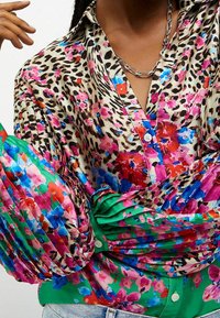 Kukkakuviollinen pusero, jossa on beigeä, vihreää ja vaaleanpunaista, laskostetut vihreät hihat ja nappikiinnitys edessä. Hopeaketjukaulakoru koristeena.