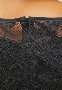 Svart spetsplagg med ett blommönster, som har en diskret dragkedjestängning bak och en slät halslinning.