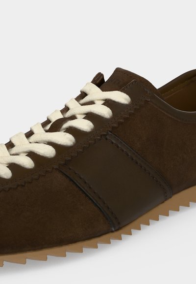 Gros plan d'une basket en daim marron avec des lacets blancs, une bande latérale en cuir foncé et une semelle en caoutchouc beige avec des bords dentelés.