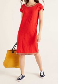 Femme portant une robe rouge longueur genou avec des bordures multicolores, des baskets bleu marine, tenant un grand sac cabas jaune moutarde avec des poignées noires.