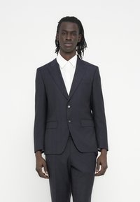 Tamsiai mėlynas kostiumas su siauru siluetu, su neišpjautais atlapais ir dviejų sagčių užsegimu. Turi šonines kišenes ir tekstūruotą audinį. Po juo balti marškiniai.