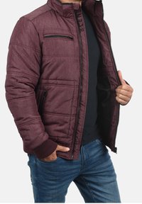 Burgunderrote Steppjacke mit Frontreißverschluss, Seitentaschen und gerippten Bündchen. Verfügt über eine strukturierte Oberfläche und einen hohen Kragen. Über einem schwarzen Hemd getragen.