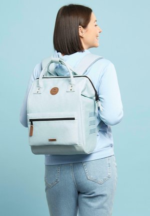 Light blue backpack with dual handles and adjustable straps, textured fabric, front zipper pocket, and leather accent logo.