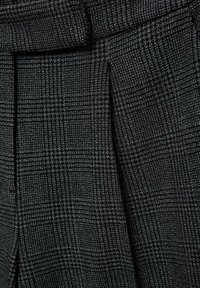 Gros plan sur un tissu gris foncé avec un petit motif à carreaux, montrant une couture et un détail de rabat.