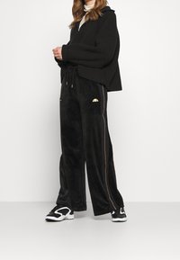 Svart ribbad huvtröja, kombinerad med svarta velourbyxor med dragkedjefickor och guldaccenter, bärs med svarta sneakers.