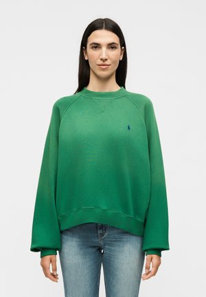 Frau mit glatten dunklen Haaren trägt grünes Sweatshirt mit langen Ärmeln und hellblaue Jeans, steht vor schlichter weißer Hintergrund.