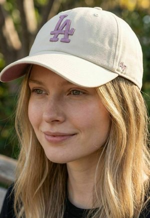 Frau mit langen blonden Haaren, die eine beige Mütze mit lila "LA"-Logo trägt, lächelt sanft, im Freien vor einem unscharfen grünen Hintergrund.