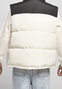 Polsterweste mit einem schwarzen oberen Teil und einem off- weißen unteren Teil. Hat einen hohen Kragen und eine glatte Textur. Getragen über hellen Jeans.