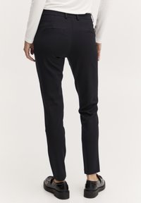 Schwarze, taillierte Hose mit schmaler Passform, glatter Textur, zwei Gesäßtaschen und mittelhohem Bund. Getragen mit schwarzen Schuhen.