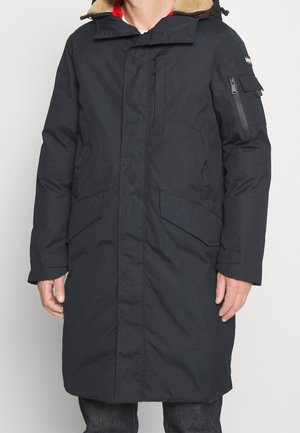 Une personne porte une longue parka d’hiver noire, avec une capuche doublée de fourrure, des poches à l’avant et une poche zippée sur la manche.