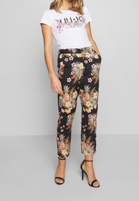 Vrouw in zwarte bloemenbroek met ananassen, een wit T-shirt met opdruk, en zwarte hoge hakken, staande tegen een effen achtergrond.