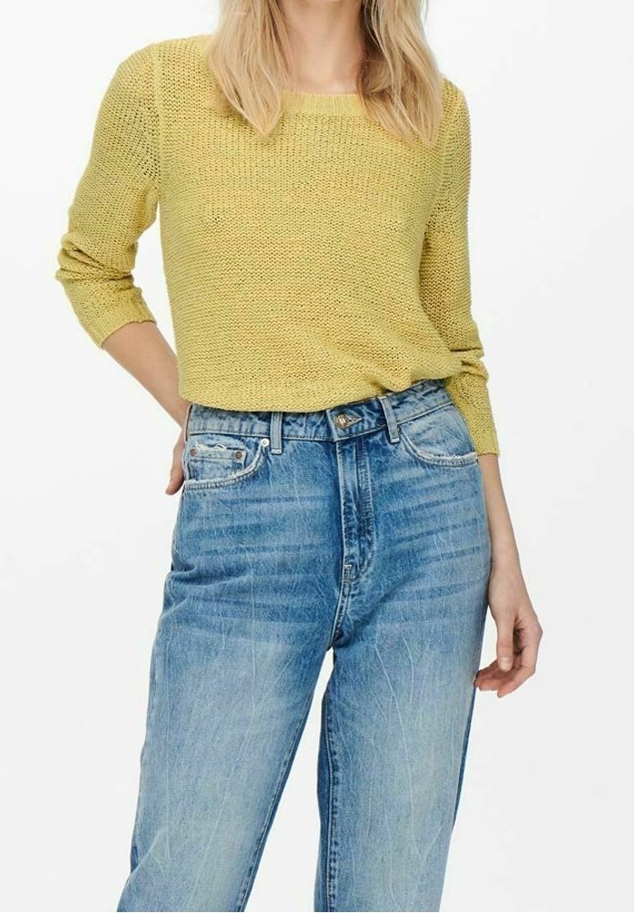 Femme portant un pull en tricot jaune et un jean bleu taille haute avec un léger délavage, debout avec une main dans la poche contre un fond blanc.