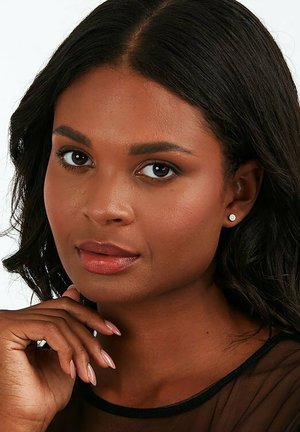 Nahaufnahme einer Person mit mittlerer brauner Haut, dunklem, welligem Haar, leichtem Make-up und kleinen Steckerohrringen, die die Hand unter das Kinn legt und ein schwarzes Oberteil trägt.