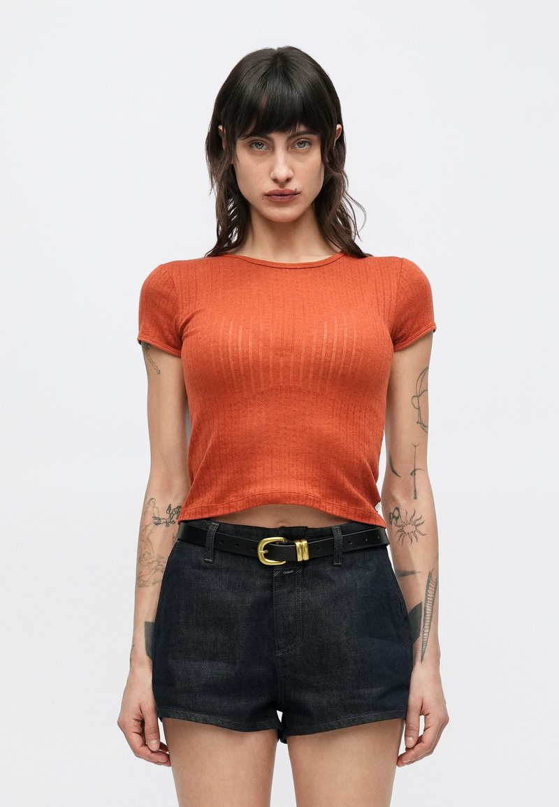 Κοντομάνικο μπλουζάκι σε σκουριά απόχρωση με ριμπ υφή, κοντά μανίκια και στρογγυλή λαιμόκοψη, συνδυασμένο με μαύρο τζιν σορτς και μαύρη ζώνη.