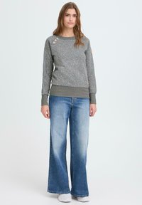 Szürke pulóver virágmintával, bordázott mandzsettákkal és három gombbal a bal vállnál. Világoskék, bőszárú farmerrel párosítva.