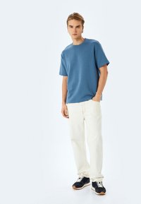 Blå kortärmad t-shirt, avslappnad passform, kombinerad med krämfärgade jeans. Svarta sneakers med vita snören och gummisula fulländar outfiten.