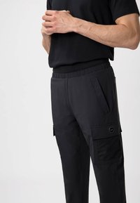Zwarte cargobroek met een elastische tailleband, voorzien van zijzakken, gaasdetails en een kenmerkende hexagonale logopatch. Vloeiende textuur.