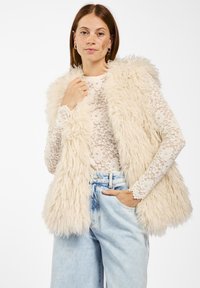 Crème faux fur vest met een zachte, gestructureerde afwerking en een capuchon, gedragen over een wit kanten shirt met lange mouwen en lichtblauwe, wijdvallende jeans.