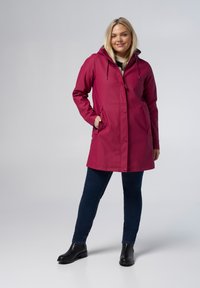 Fuchsia waterdichte jas met capuchon, drukknoopsluiting en zijzakken, gecombineerd met donkere jeans en zwarte enkellaarsjes. Gladde textuur.