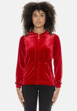 Frau mit lockigem Haar, die einen glänzenden roten Kapuzenpullover mit Reißverschluss und Fronttaschen sowie schwarze Hosen trägt, steht vor einem schlichten hellen Hintergrund.