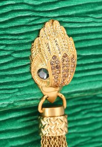 Zawieszka w złotym odcieniu z motywami liści i oka, ozdobiona przezroczystymi i ciemnymi kryształkami, zawieszona na plecionym łańcuszku. Tło zielone.