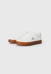 Witte lage sneakers met bruine gumzolen, veters en een groen "ck"-logo aan de zijkant, tegen een eenvoudige witte achtergrond.