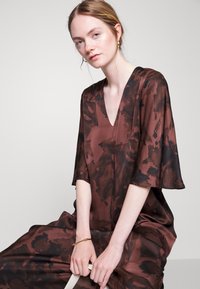 Vestido castanho com padrão, decote em V e mangas largas. Feito de um tecido suave, drapeia-se de forma solta sobre o corpo, apresentando acentos escuros subtis.