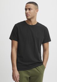 Camiseta de algodón negra con cuello redondo y manga corta. Presenta un sutil logo de la marca en la parte superior izquierda del pecho. Textura suave.