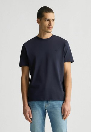 T-shirt in cotone blu navy con collo rotondo, maniche corte e un logo discreto sul petto, abbinata a jeans blu chiaro.