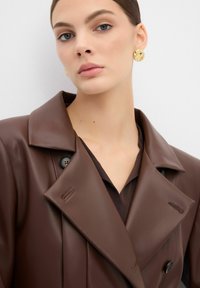Bruine faux leren jas met een brede kraag, voorzien van decoratieve stiksels en zwarte knopen. Draagt over een donker shirt. Gouden oorbellen zichtbaar.