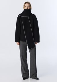 Abrigo negro de gran tamaño con cuello alto y costuras en contraste gris. Combinado con pantalones grises de pierna ancha y zapatos negros con punta. 