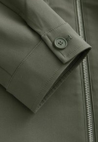 Olivgrüne Jacke mit einer glatten Stofftextur, einem Reißverschluss vorne und einem knöpfbaren Ärmeldetails.
