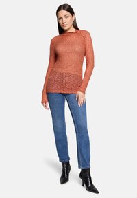 Roestkleurige kanten top met lange mouwen, een aansluitend ontwerp, gelaagde textuur en hoge hals, gecombineerd met blauwe jeans en zwarte hakken.