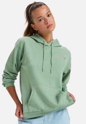 Jeune femme avec une queue de cheval portant un sweat à capuche vert clair avec le logo "ROXY", posant les mains près des poches contre un fond blanc.