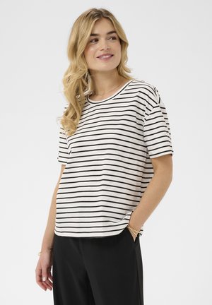 Blonde Frau trägt ein weißes kurzärmeliges Shirt mit schwarzen horizontalen Streifen und schwarze Hose, lächelt und blickt zur Seite.
