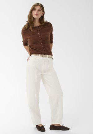 Un pull en maille côtelée marron à boutons, un pantalon blanc à jambes larges avec des coutures latérales, une ceinture beige et des chaussures en daim marron foncé, le tout sur fond blanc.