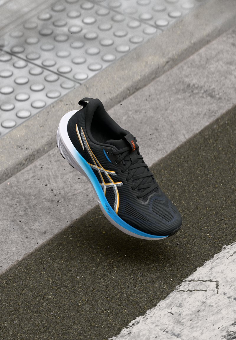 Zapatilla deportiva negra con parte superior texturizada, entresuela azul y blanca, y acentos en naranja y blanco en contraste. Cuenta con cordones y una puntera redondeada.