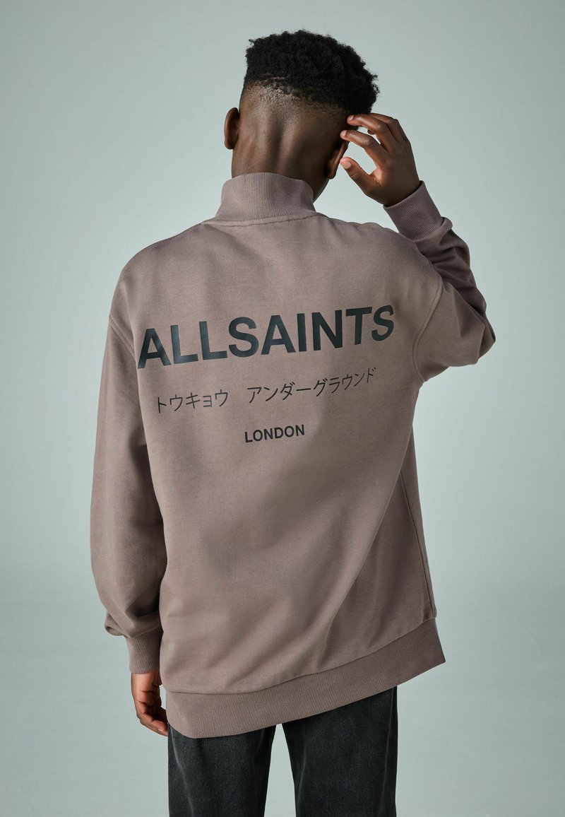 Felpa oversize marrone con colletto alto, caratterizzata da testo stampato in nero sul retro e polsini a coste. Materiale morbido in misto cotone.