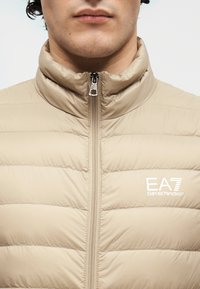 Casaco bege acolchoado com gola alta, apresentando costura horizontal, fecho de correr completo e logótipo EA7 Emporio Armani branco no peito.