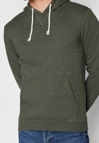 Uomo che indossa un felpa oliva con cappuccio, caratterizzata da lacci bianchi e tasca frontale, abbinata a jeans blu, su uno sfondo neutro.