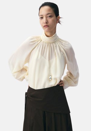 Kobieta o ciemnych włosach związanych do tyłu nosi prześwitującą kremową bluzkę z wysokim kołnierzem i czarną warstwową spódnicę, z rękami na biodrach, neutralnym wyrazem twarzy.