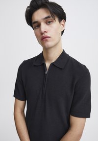 Polo scurt cu mâneci negre, cu guler cu fermoar, dintr-un material texturat, cu un design ajustat. Detaliile subtile includ margini ribbate și umeri fără cusături.