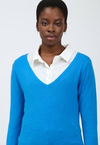 Helderblauwe v-hals trui van zachte stof, gedragen over een witte blouse met kraag. Beschikt over lange mouwen en een aansluitende silhouette.