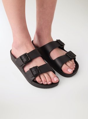 Piedi che indossano sandali neri tipo slide con due cinturini regolabili con fibbia su uno sfondo chiaro.