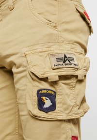 Beżowe spodnie cargo z kieszenią i naszywką "Alpha Industries" oraz naszywką "Airborne" z głową orła na niebieskim tle.