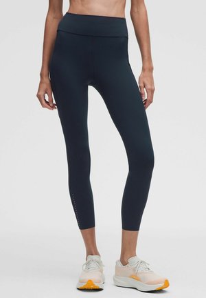 Kvinde iført sorte højtaljede korte leggings og hvide sportssko med orange detaljer, stående mod en ensfarvet baggrund.